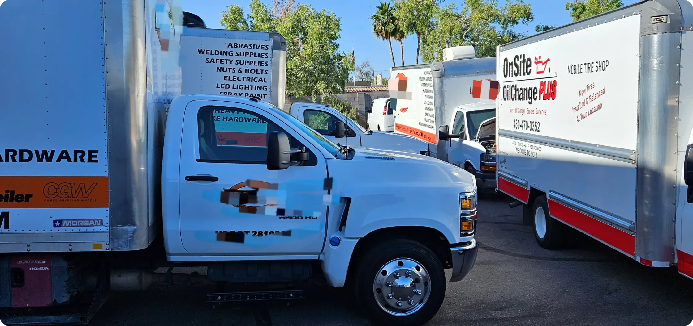 Several white utility trucks with business logos, including a mobile auto repair service, are parked closely together in a lot on a sunny day, surrounded by trees and palm trees in the background.