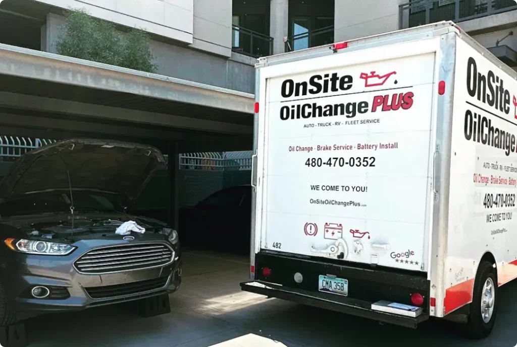 A gray sedan with its hood open is parked next to a white OnSite OilChange Plus mobile auto repair service truck in a covered parking area near a building.