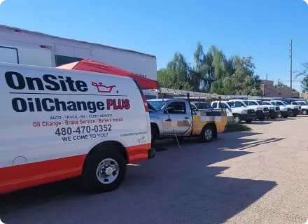 A white and red OnSite OilChange PLUS van, offering mobile auto repair services, is parked beside several white utility trucks in a dirt lot under a clear blue sky. Trees and a building can be seen in the background.