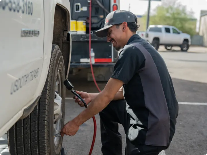 A mechanic with a dark shirt kneels beside a white truck, smiling as he uses an air pressure gauge to check or inflate a tire—showcasing mobile auto repair with vehicles and equipment visible in the background.