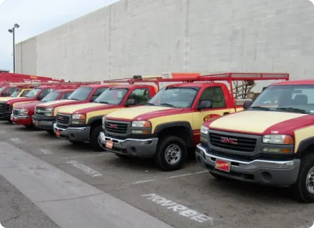 A row of red and yellow utility pickup trucks, some equipped for mobile auto repair with ladders mounted on top, are parked in designated spaces next to a large white concrete wall.