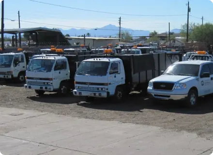 A row of white utility trucks and a mobile auto repair pickup truck with orange lights on top are parked in a dirt lot, with industrial buildings and mountains visible in the background.