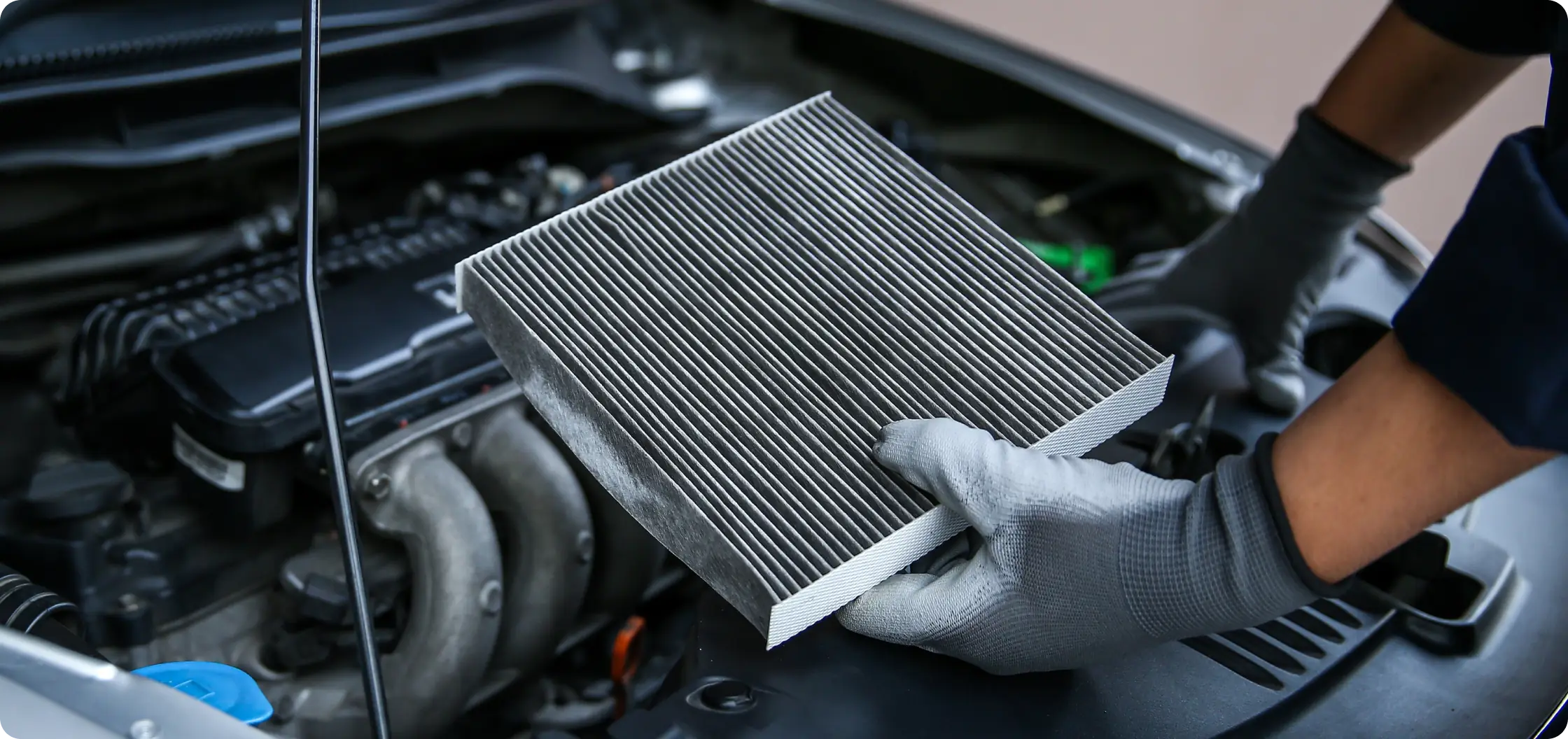 A person wearing gray gloves holds a clean rectangular car air filter above an open engine bay, preparing to replace or inspect it.