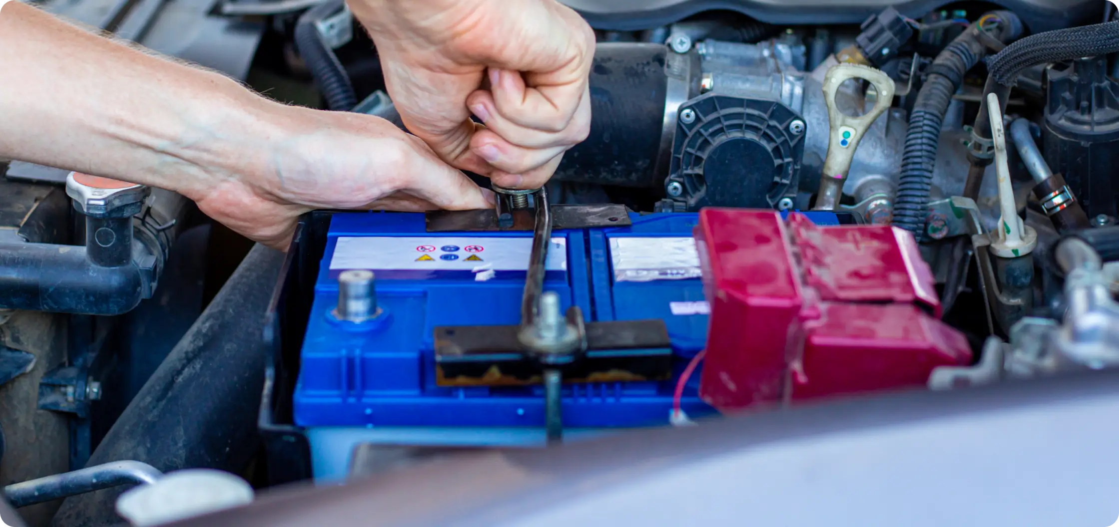 A person’s hands use a wrench to tighten a bolt on a car battery under the hood, with engine components visible in the background.