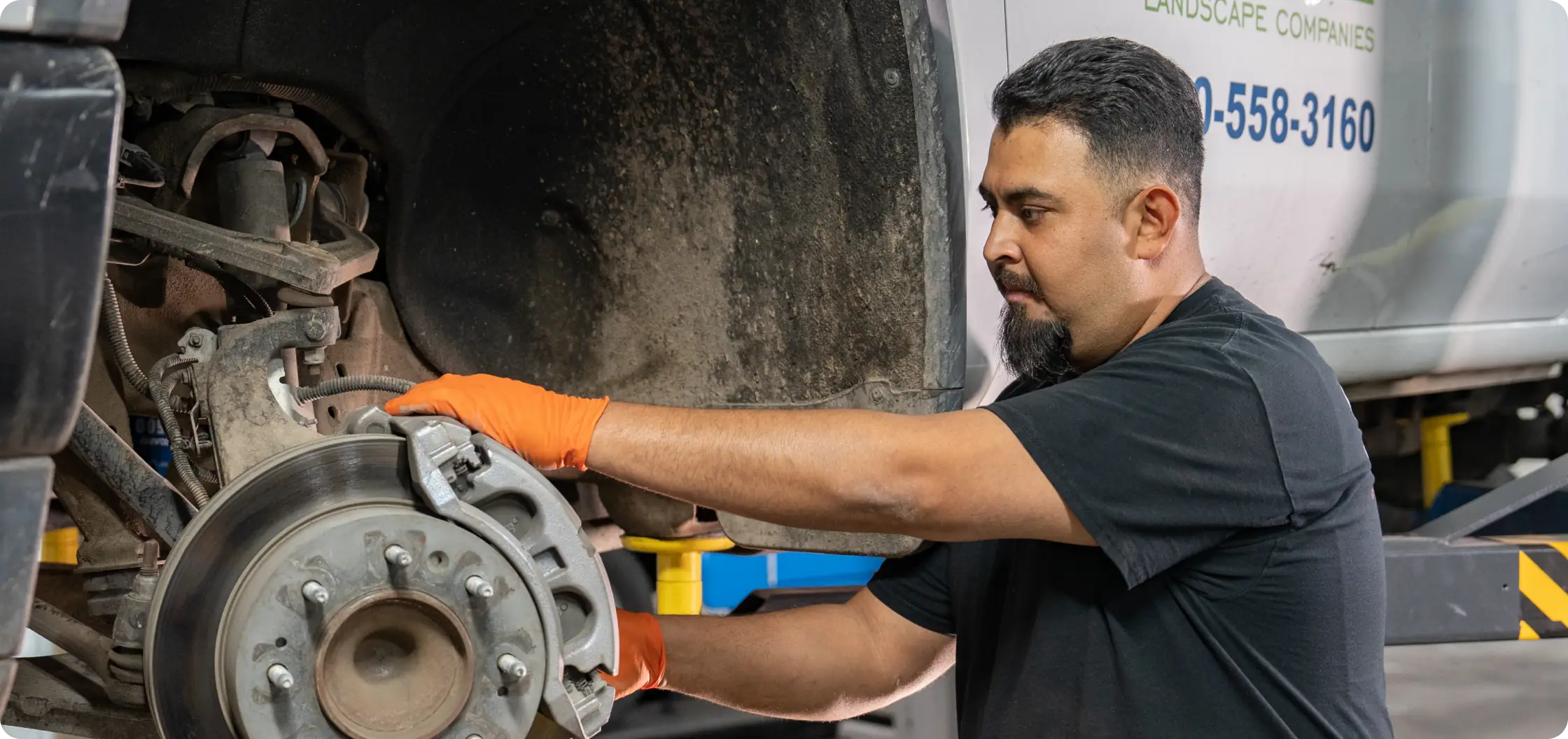 A mechanic wearing orange gloves works on the exposed brake system of a vehicle, inspecting or repairing parts inside the wheel well in a garage or workshop.