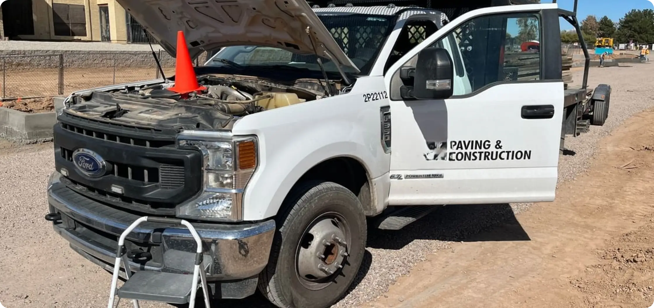 A white construction truck with its hood open and a traffic cone placed on the engine is parked on a gravel lot. The drivers door is open, and a small step ladder is positioned in front of the truck.