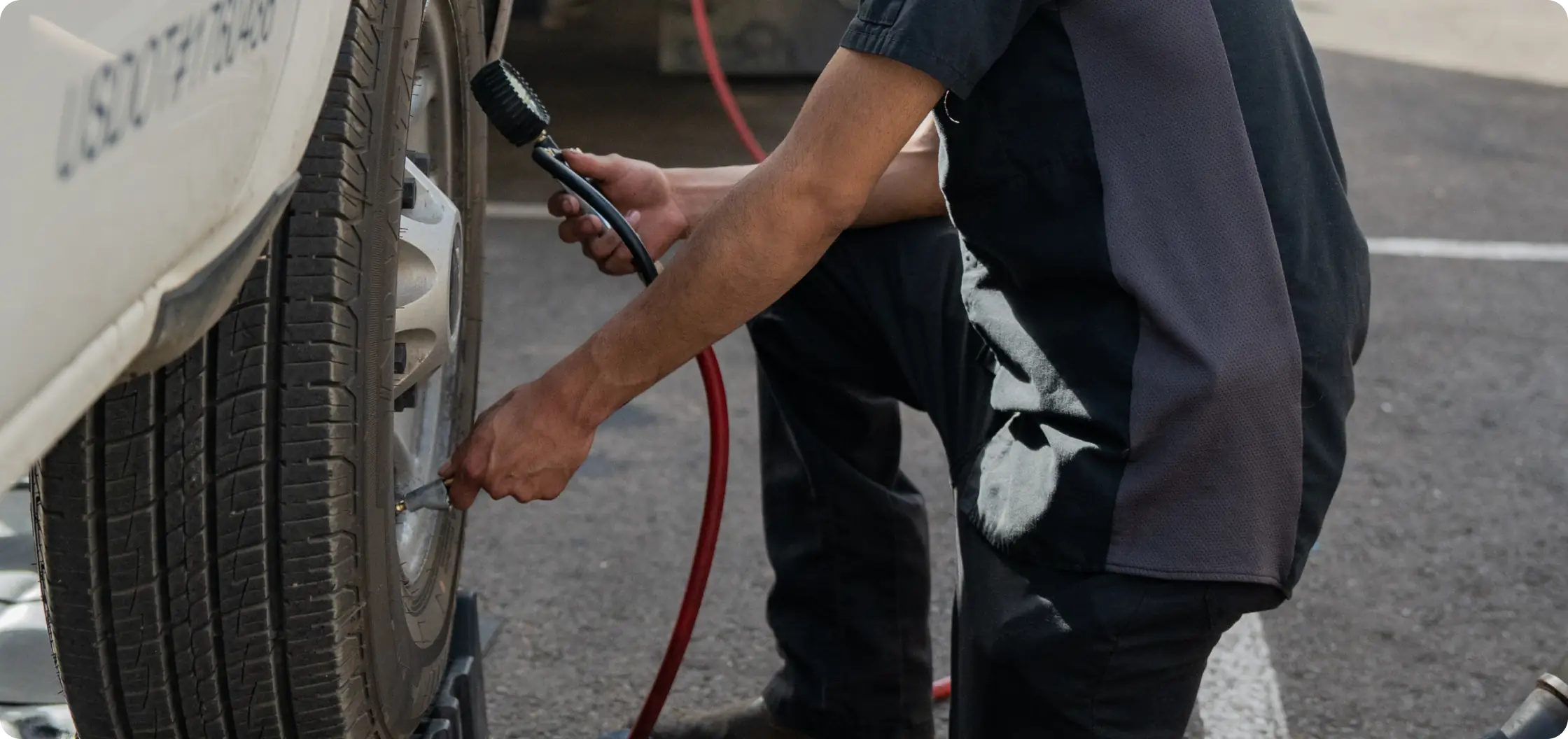 A person in work clothes is kneeling next to a car tire, using an air hose to inflate it. The car is parked on a paved surface, and the red air hose is visible on the ground.