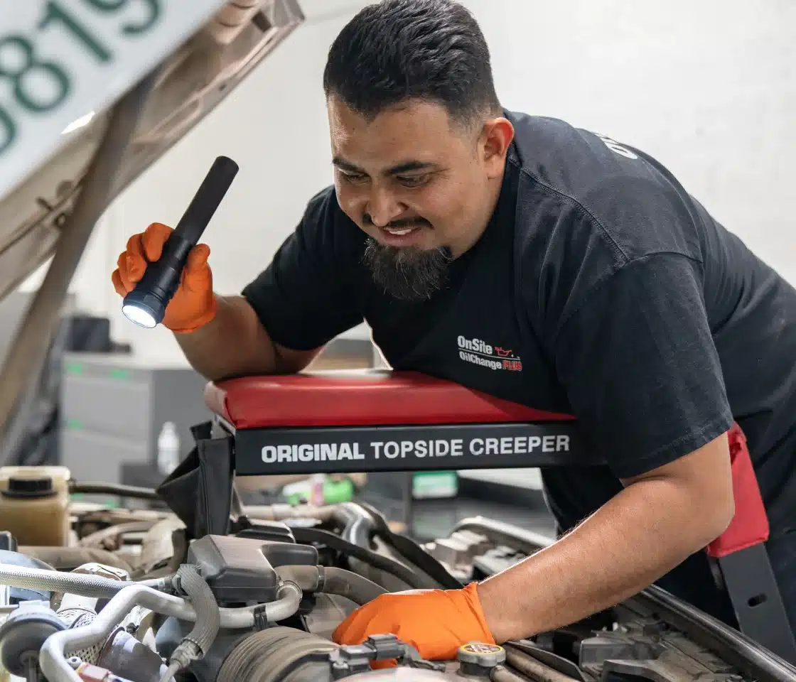 A mechanic wearing orange gloves uses a flashlight while inspecting an engine. He is leaning on a padded support labeled ORIGINAL TOPSIDE CREEPER and appears focused on his work.