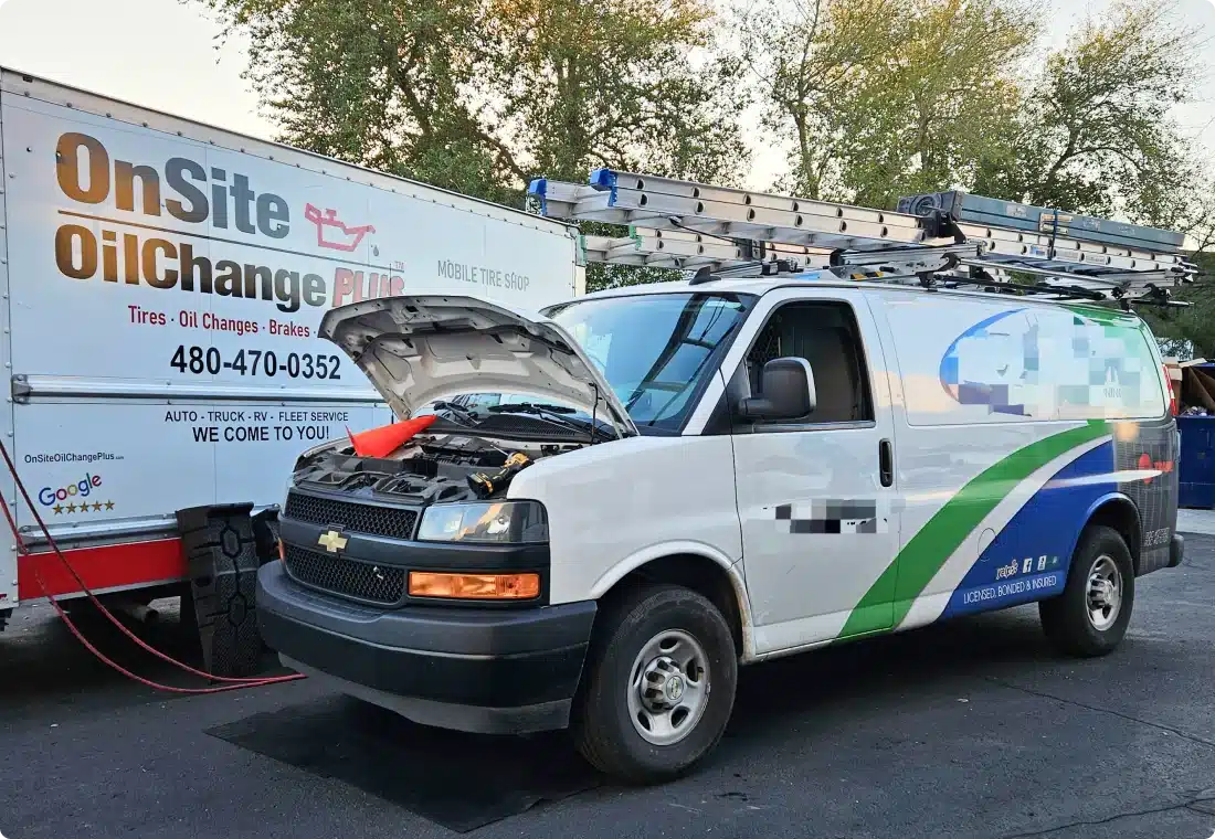 A white service van with its hood open is receiving a jump start from an OnSite OilChange truck parked nearby. Both vehicles are in a parking lot with trees in the background.