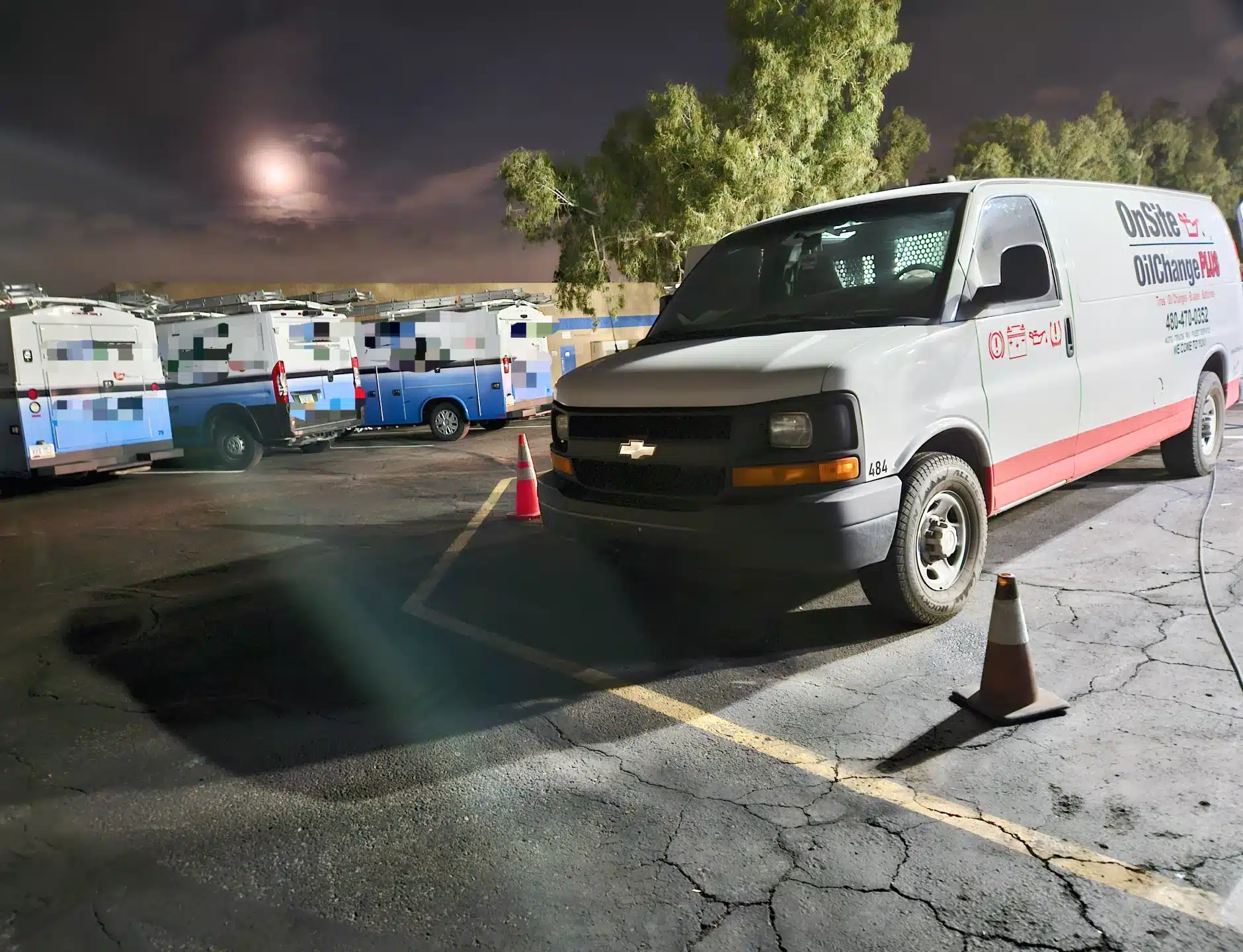 A white mobile service van with OnSite OilChange branding is parked in a lot at night near orange cones. Several other service vehicles are parked in the background under a cloudy sky with a full moon.