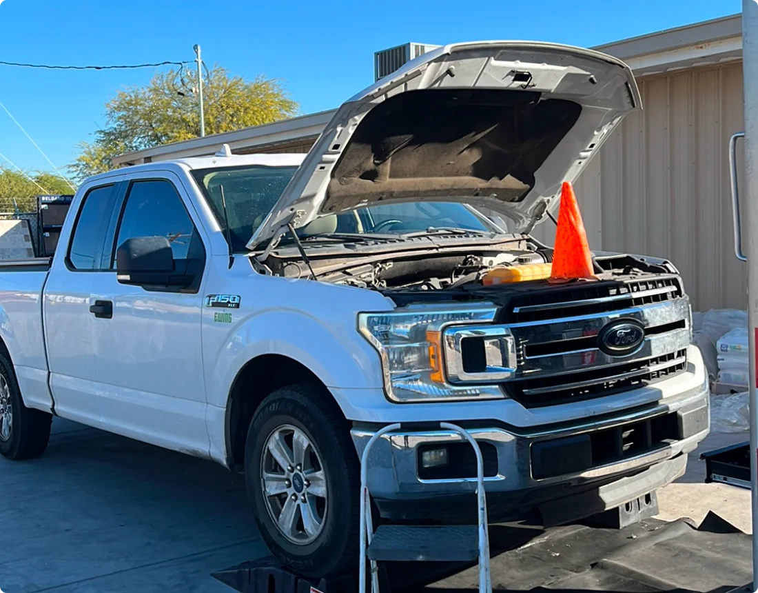 A white Ford F-150 pickup truck with its hood open is parked on a platform. An orange safety cone is placed on the front grille. A beige building and trees are visible in the background.