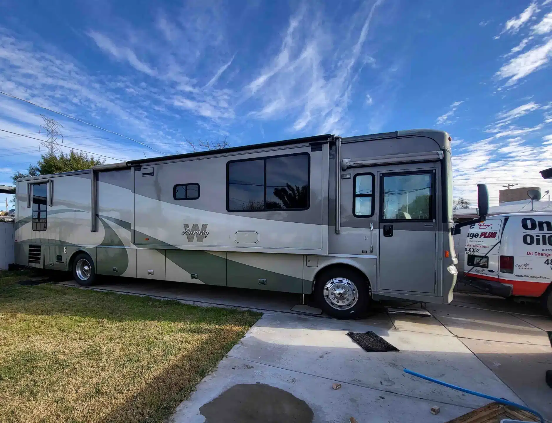 A large, silver and gray motorhome with tinted windows is parked on a driveway next to a grassy yard under a blue sky with scattered clouds. Another vehicle is partially visible on the right.