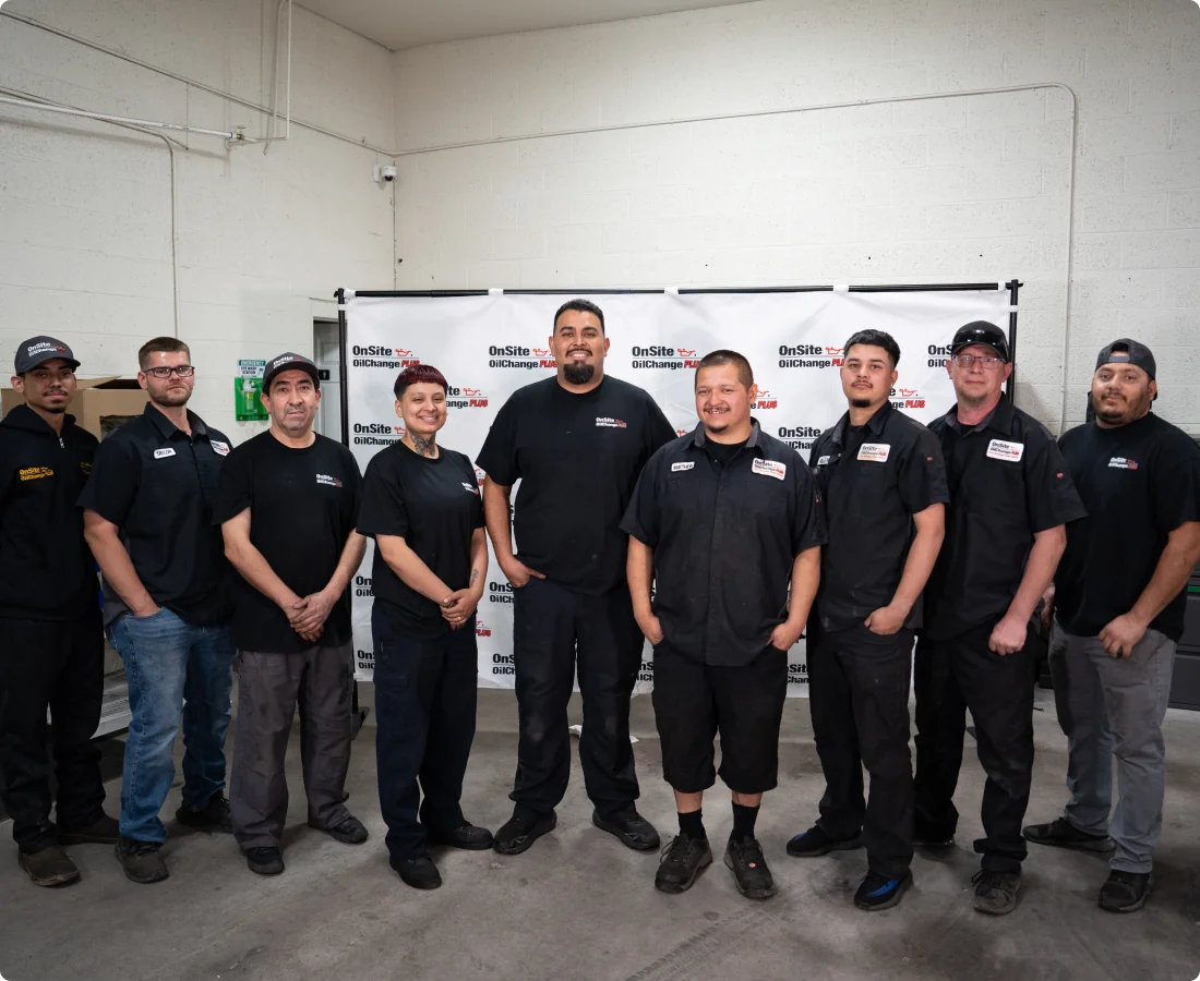 A group of ten people in black work uniforms stands together and smiles for a group photo in front of a white backdrop with company logos, showcasing what teamwork is all about inside an industrial workspace.