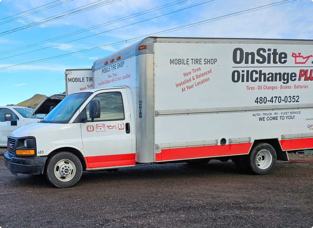 A white and red box truck with OnSite Oil Change Plus and contact information advertising mobile tire and oil change services, parked on a dirt lot with another service vehicle partially visible behind it.