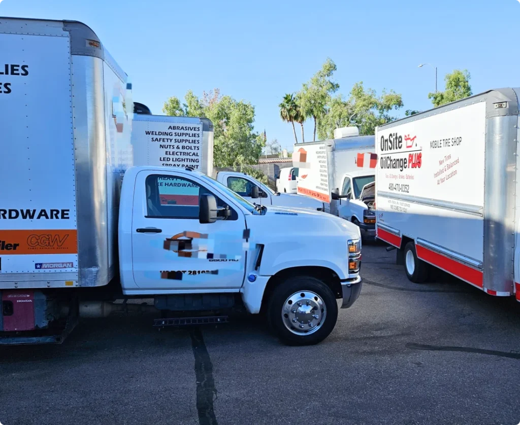 Several white utility trucks with company logos and equipment are parked in a lot on a sunny day, surrounded by trees and clear blue sky, ready for routine tasks like an oil change.